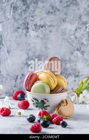 Varietà di colorati macaron dolci francesi con diversi macaron ripieni serviti in tazza da tè d'epoca con fiori di primavera e. frutti di bosco freschi Foto Stock