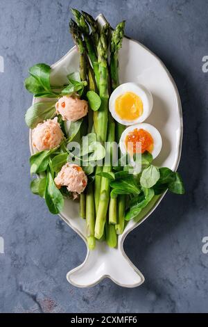Asparagi verdi cotti con uova sode a metà, insalata, caviale rosso e pate di salmone serviti su piatto a forma di pesce bianco su sfondo metallico grigio di consistenza blu Foto Stock