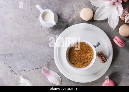 Tazza bianca di caffè nero, servita su piattino bianco con brocca di panna, biscotti macaroon e ramo di fiori magnolia su sfondo grigio Foto Stock