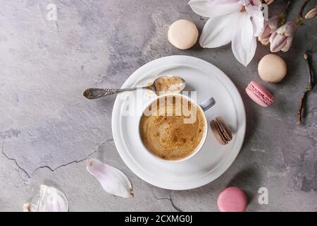 Tazza bianca di caffè nero, servita su piattino bianco con biscotti macaroon, cucchiaio e ramo di fiore magnolia su fondo grigio. Piatto Foto Stock