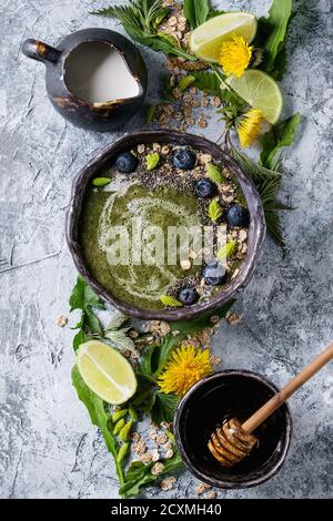 Verde primavera ortica e tarassaco ciotola smoothie servita con calce, fiori gialli, foglie giovani, fiocchi di avena, semi di Chia, mirtilli, la crema di latte e miele o Foto Stock