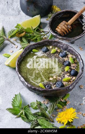 Verde primavera ortica e tarassaco ciotola smoothie servita con calce, fiori gialli, foglie giovani, fiocchi di avena, semi di Chia, mirtilli, la crema di latte e miele o Foto Stock