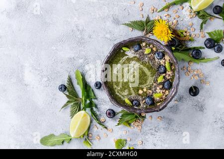 Verde primavera ortica e tarassaco ciotola smoothie servita con calce, fiori gialli, foglie giovani, fiocchi di avena, chia semi, mirtilli, crema su grigio b Foto Stock