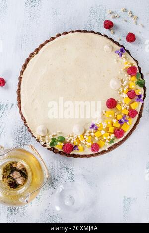 In casa torta al cioccolato decorate da mango, lamponi, menta, il riso soffiato e fiori commestibili servita con vetro teiera su bianco texture backg in legno Foto Stock