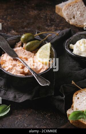 Ciotola nero di pâté di salmone con caviale rosso serviti con burro, pane affettato, capperi, vintage coltello e erbe sul lino tessile igienico su texture marrone Foto Stock