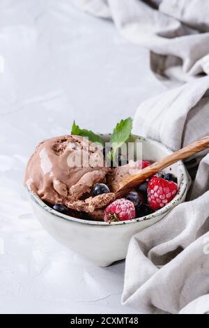 Sciogliendo gelato artigianale al cioccolato con mirtillo di frutti di bosco surgelati, lampone, menta servita in ciotola bianca con cucchiaio di legno d'oliva e lino tessile Foto Stock