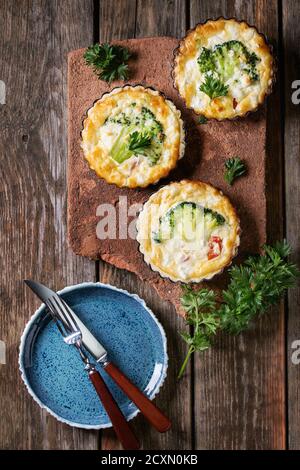 Cotto in casa quiche torta in mini metallo forma servita con verdure fresche, piastra e posate sulla scheda di terracotta sul vecchio plank sullo sfondo di legno. Lay piatto Foto Stock