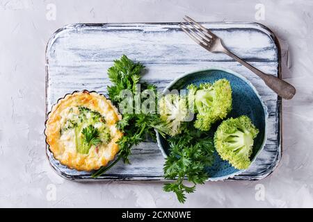 Cotto in casa verdure broccoli quiche torta in mini metallo forma servita con verdure fresche, piastra forcella su bianco che serve di bordo su cemento grigio backgro Foto Stock