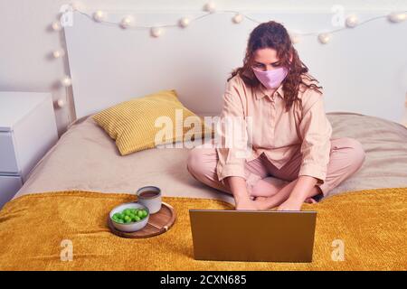 Donna rossa in maschera medica lavora su computer portatile in letto giallo. Ragazza lavora al computer online durante l'isolamento dovuto virus influenzale, concetto Foto Stock