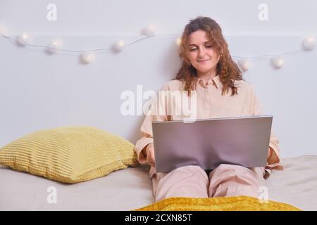 Una donna sorridente si siede con un computer portatile e lavora su un letto bianco. Ragazza felice con un computer in camera da letto, primo piano Foto Stock