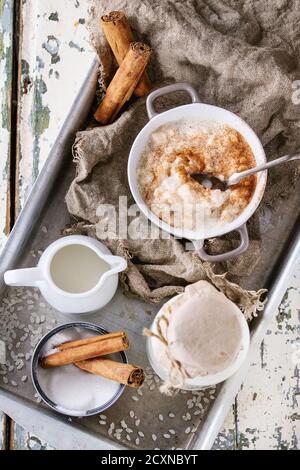 Ingredienti per fare budino di riso. Riso crudo, zucchero, bastoncini di cannella, latte, caraffa di panna e pentola di budino da cucina su vassoio di alluminio su bianco Foto Stock