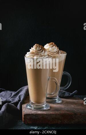 Zucca piccante di latte con panna montata e cannella in due bicchieri in piedi sul bordo di argilla con altri tessili sfondo scuro. Dolce calda bevanda di caffè Foto Stock