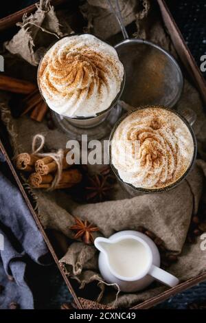 Latte speziato di zucca con panna montata e cannella in due bicchieri in scatola di legno con tessuto, brocca di panna e spezie altro sfondo scuro. Foto Stock