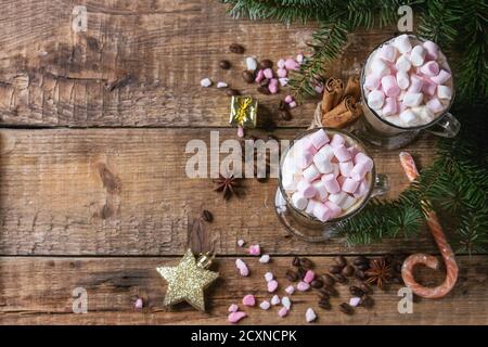 Due bicchieri di caffè latte con marshmallow rosa con decorazioni natalizie, zucchero rosa, caramelle, spezie, chicchi di caffè e abeti su tessuto di legno backgro Foto Stock