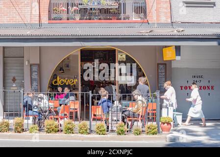 La gente siede socialmente distanziata fuori di un caffè mentre una donna che indossa una maschera cammina attraverso il sobborgo di Sydney di Annandale, nuovo Galles del Sud, Australia Foto Stock