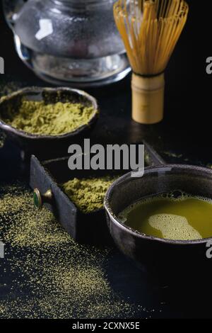 Il tè verde Matcha polvere e bevanda calda in nero ciotole e scatola di legno in piedi con vetro teiera, bambù strumenti tradizionali cucchiaio, frusta nel vassoio di terracotta Foto Stock