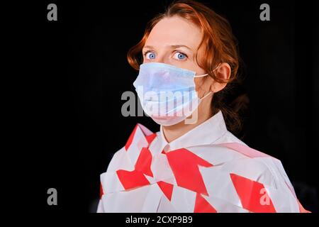 Donna legata con occhi spaventati in maschera medica su sfondo nero, concetto. Primo piano della ragazza rossa durante la quarantena del coronavirus Foto Stock