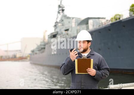 Ufficiale del ponte marino che parla da radio walkie talkie e in piedi vicino nave con documenti in mani. Foto Stock