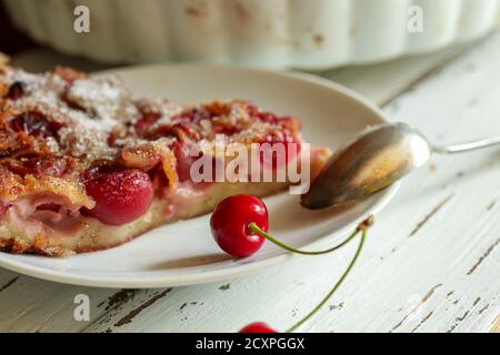 Una fetta di dessert di clafoutis fatto in casa - tradizionale torta di ciliegie francesi, ciliegie fresche e un cucchiaino da tè su un piatto bianco su un tavolo di legno bianco, c. Foto Stock