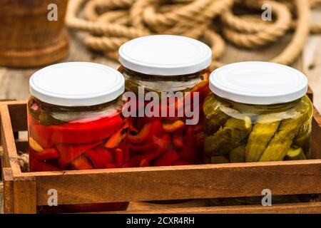 Cassa di legno con vasi di vetro con peperoni rossi decapati e cetrioli decapati (sottaceti) isolati. Vasetti con varietà di verdure sottaceto. Conserva Foto Stock