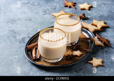 Cocktail al latte di Natale eggnog con cannella, servito in due bicchieri con biscotti allo zucchero a forma di stella di shortbread di dimensioni diverse rispetto allo sfondo a struttura blu Foto Stock