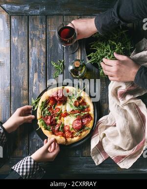 Tenendo le mani a fette di pizza fatta in casa con formaggio e bresaola, servita su piastra nera con rucola fresca, olio di oliva, un bicchiere di vino rosso e asciugamano da cucina Foto Stock