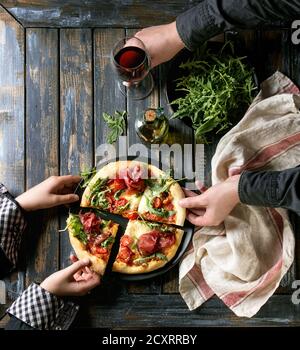 Tenendo le mani a fette di pizza fatta in casa con formaggio e bresaola, servita su piastra nera con rucola fresca, olio di oliva, un bicchiere di vino rosso e asciugamano da cucina Foto Stock