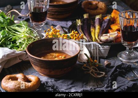 Holiday table decoration setting with bowls of hot soup, baking pumpkin, carrot, garlic, fresh coriander, pretzels bread, red wine, berries over woode Stock Photo