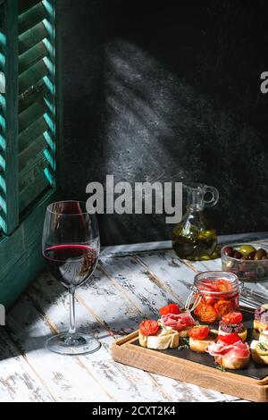 Le tapas o bruschetta varietà. Fette di pane con prosciutto, pomodori secchi, olio di oliva, olive, pepe su ardesia scheda di legno con un bicchiere di vino rosso al di sopra Foto Stock