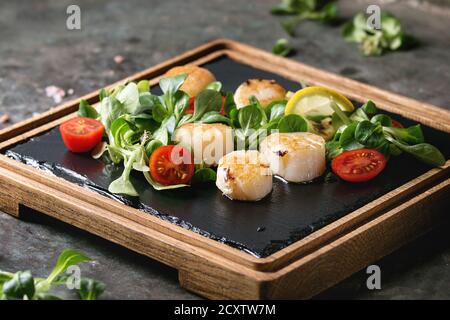 Cappesante fritte con limone, pomodorini e insalata verde servita su legno nero ardesia scheda di servizio sopra il vecchio metallo scuro dello sfondo. Close up Foto Stock