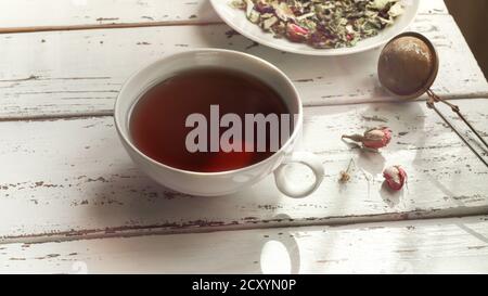 Tè alle erbe - balsamo al limone, tiglio, verbena, boccioli di rosa, lavanda in un piatto bianco e in una tazza bianca, e cucchiaio infusore di tè su un tavolo di legno chiaro Foto Stock
