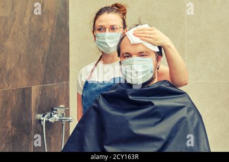 Una donna parrucchiere in una maschera medica è venuto a fare i suoi capelli per un uomo cliente. Concetto di recupero e di ritorno alla vita normale dopo l'epidem del coronavirus Foto Stock