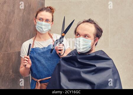 Moglie parrucchiere in maschera medica fa taglio di capelli al marito con forbici da giardino, concetto divertente Foto Stock