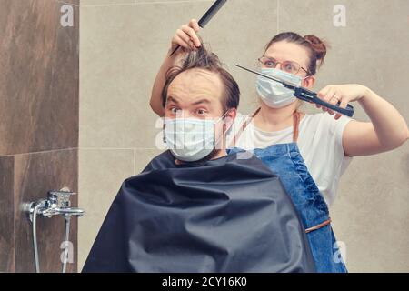 Donna parrucchiere in maschera medica fa taglio di capelli per l'uomo con grandi tagliatori, concetto divertente Foto Stock