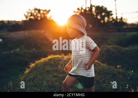 Ragazzo piccolo che gioca in un campo mentre indossa cappello e. il sole lo riscalda in una serata estiva Foto Stock