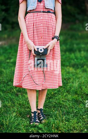 Ragazza senza volto in abiti retrò tiene una macchina fotografica vintage su uno sfondo di erba verde. Foto Stock