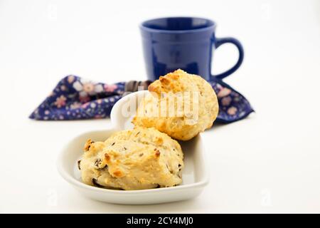 Cibo di comfort con biscotti caldi a goccia di uva passa in primo piano con tazza blu scuro e tovagliolo fiorito dietro su sfondo bianco Foto Stock