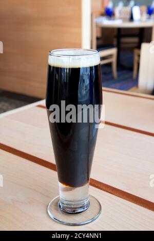 Birra scura con schiuma sopra in vetro alto sopra il tavolo sembra freddo e allettante Foto Stock