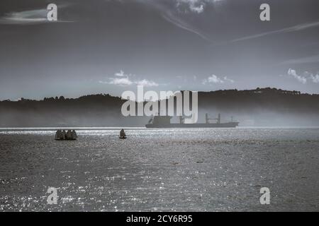 Seascape con sagome di enorme nave da carico o una chiatta e un gruppo di piccole barche a vela sportive, sparato in chiave scura in una giornata di sole con una costa collinare in Foto Stock