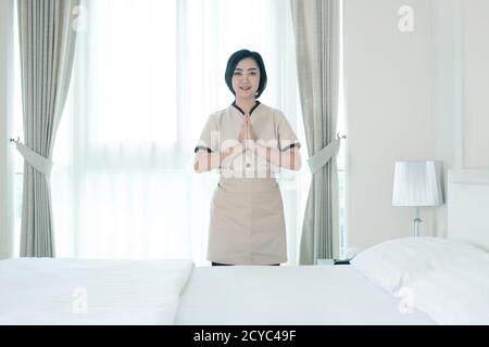 La giovane cameriera asiatica alza una mano per pagare rispetto nella camera d'albergo, guarda la macchina fotografica Foto Stock