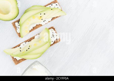 Fresco estivo salutare antipasto di pane di segale intero con fette di avocado, olio d'oliva, semi di sesamo nero, formaggio cremoso su tavola di legno bianco, Foto Stock