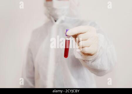 un tecnico di laboratorio in una tuta protettiva e maschera contiene una fiala piena di sangue per l'analisi di virus o malattie. Test del DNA. Mano che tiene un sangue Foto Stock