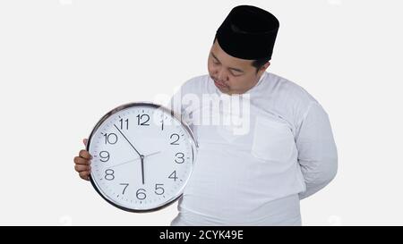 musulmano asiatico con sovrappeso in piedi e tenere grande orologio rotondo in attesa di pausa digiuno. islam ragazzo grasso non può aspettare per mussulmano pausa digiuno conc Foto Stock