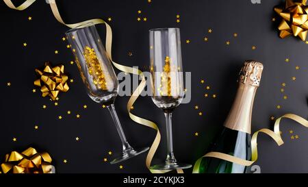 Concetto di festa di Capodanno. Sfondo natalizio nero con bottiglia di champagne, bicchieri da bibita, stelle di confetti dorati. Disposizione piatta, vista dall'alto. Foto Stock