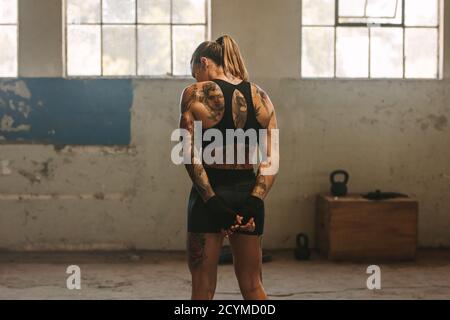 Vista posteriore di una femmina in abbigliamento sportivo e guanti da boxe all'interno del vecchio magazzino. Pugile tatuato donna in procinto di iniziare all'interno del magazzino abbandonato. Foto Stock