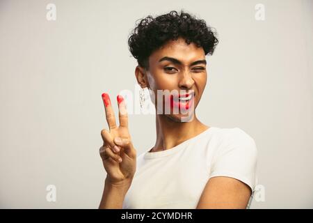 Uomo androgino con rossetto sbavato che mostra un segno di pace. Uomo transgender con rossetto che guarda la fotocamera. Foto Stock