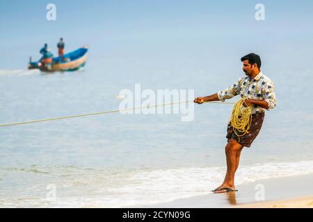 Pescatore vestito in un lungi che tira grande rete di senna spiaggia a riva alla trafficata, popolare spiaggia di Marari; Mararikulam, Alappuzha (Alleppey), Kerala, India Foto Stock