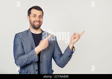 Giovane uomo d'affari con barba in giacca, sorridendo e guardando la macchina fotografica, puntando con le mani e le dita a lato. Ritratto di un uomo su sfondo grigio. Foto Stock