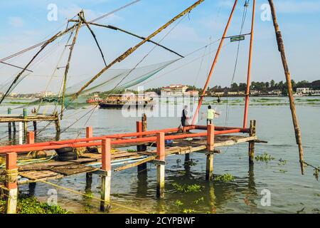 Vecchie reti da pesca cinesi, una barca turistica e rete di fusione uomo sul lungomare di Vypin Island, con Fort Cochin oltre; Fort Vypin, Kochi, Kerala, India Foto Stock