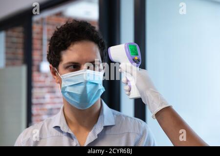 Razza mista donna e uomo caucasico che lavora in un ufficio casual, indossando maschera facciale, una donna che prende la sua temperatura. La distanza sociale sul posto di lavoro d Foto Stock